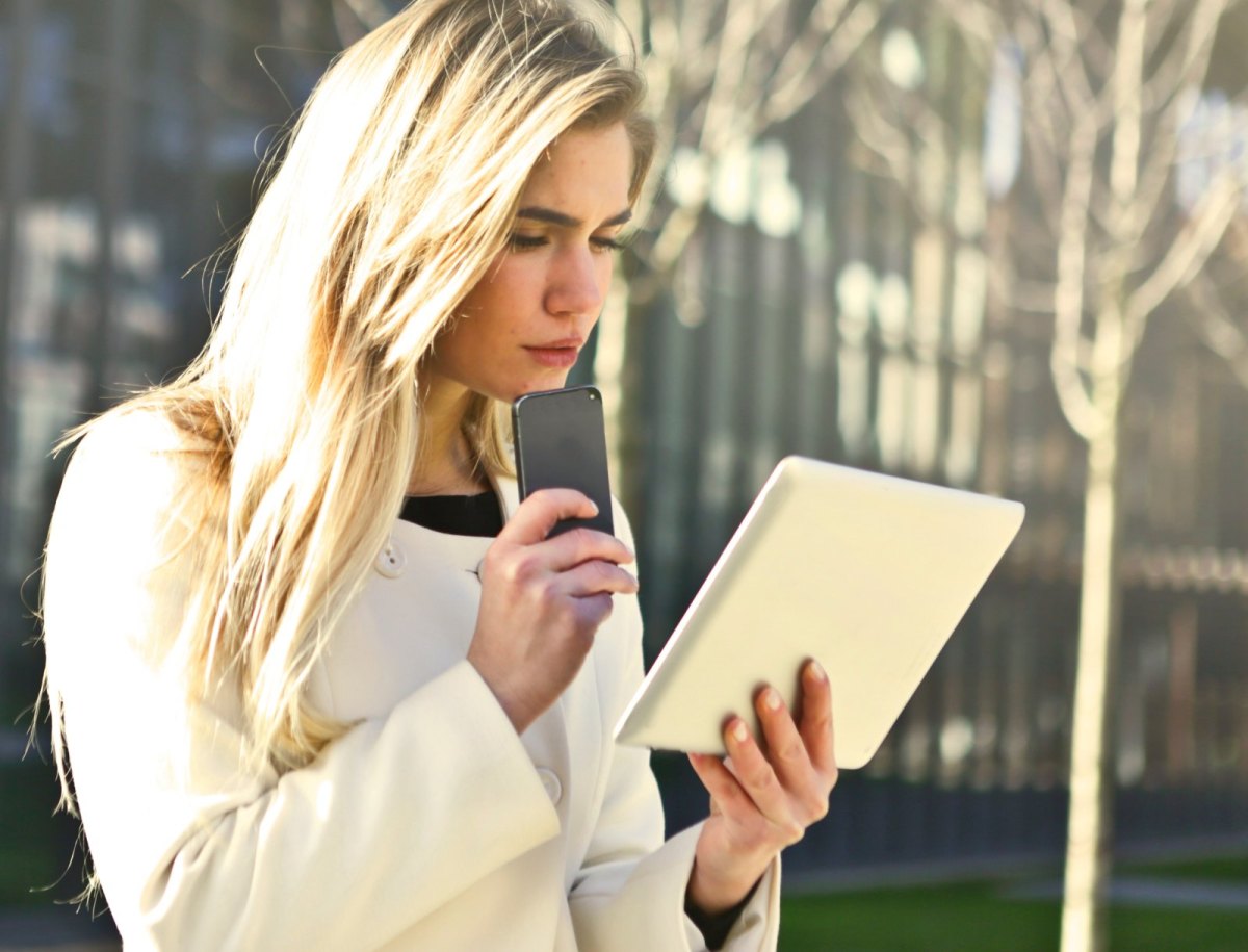 Junge Frau steht mit Smartphone und Tablet vor einem Gebäude und schaut verdutzt.