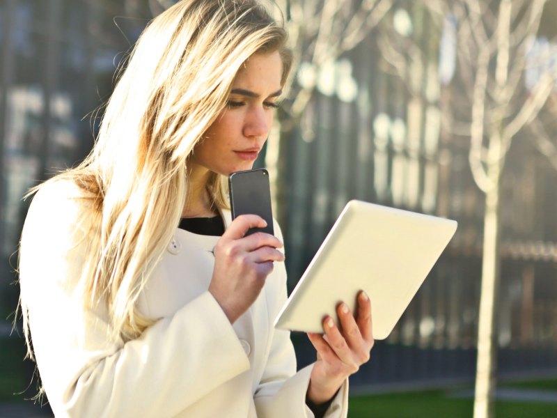 Junge Frau steht mit Smartphone und Tablet vor einem Gebäude und schaut verdutzt.