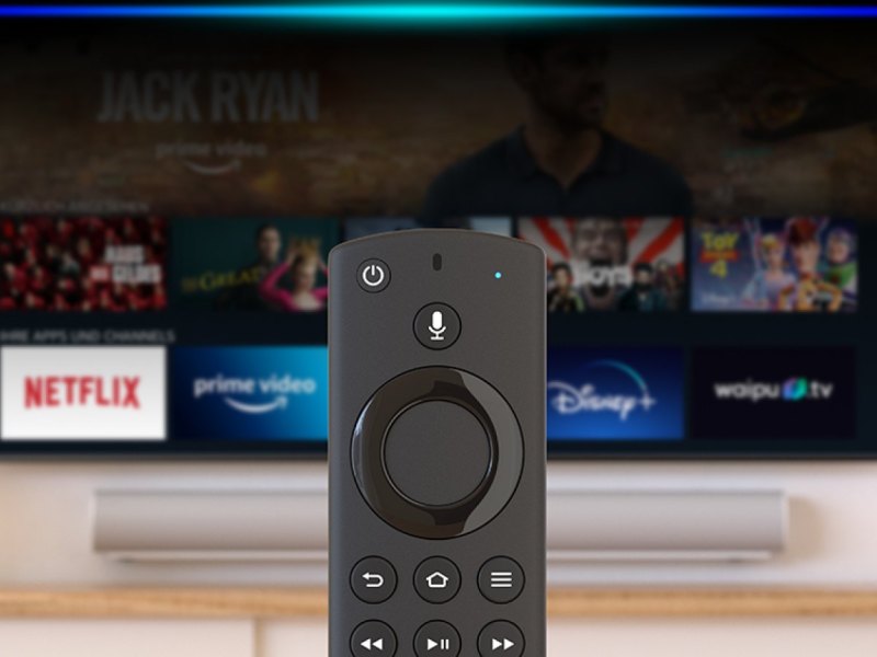 Ein Amazon Fire TV Stick im Vordergrund. Dahinter ein Fernseher mit der Amazon Fire TV-App.