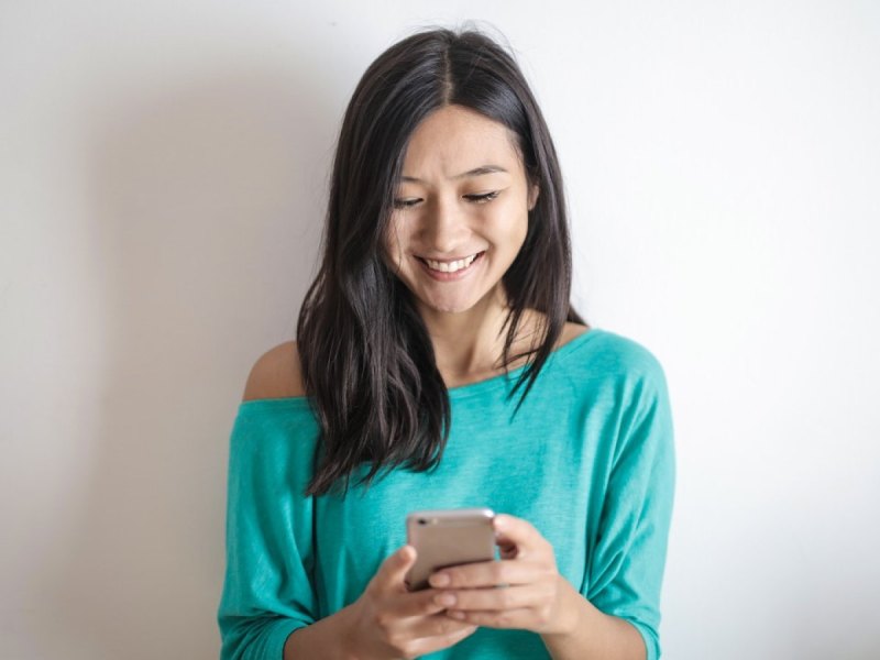 Eine Frau steht vor einer Wand und schaut lächelnd auf ihr Smartphone.