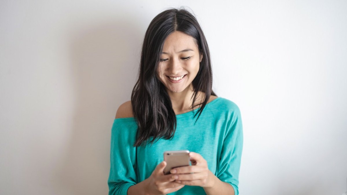 Eine Frau steht vor einer Wand und schaut lächelnd auf ihr Smartphone.