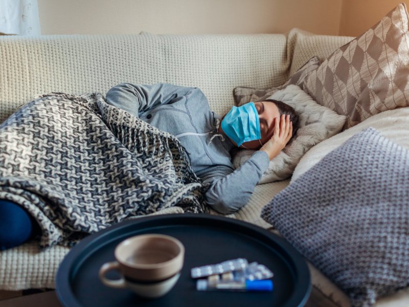 Frau liegt mit Maske krank auf dem Sofa.