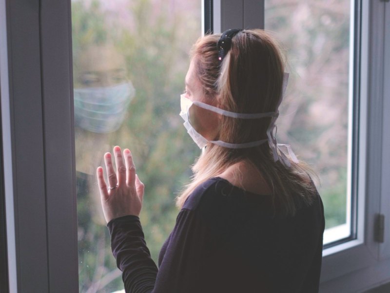 Frau mit Mundschutz schaut aus dem Fenster.