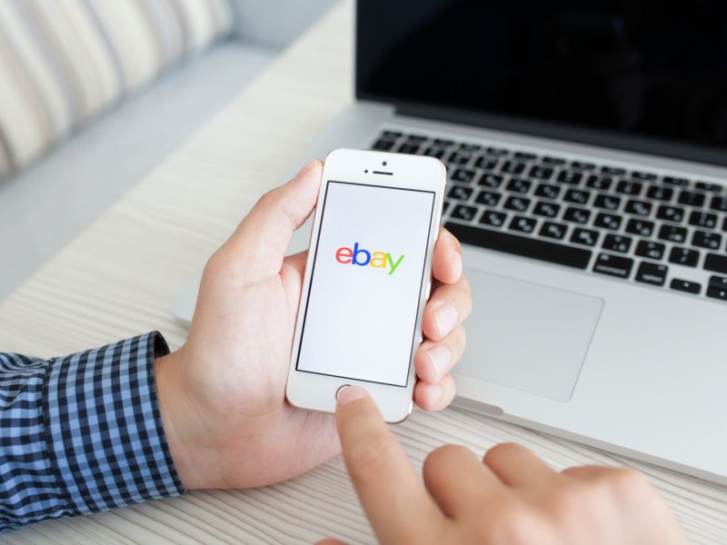 Mensch hält Handy mit eBay-Logo in der Hand.