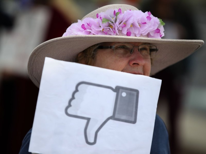 Eine Frau mit Hut hält ein Papierschild mit einem Facebook-Dislike-Button hoch.