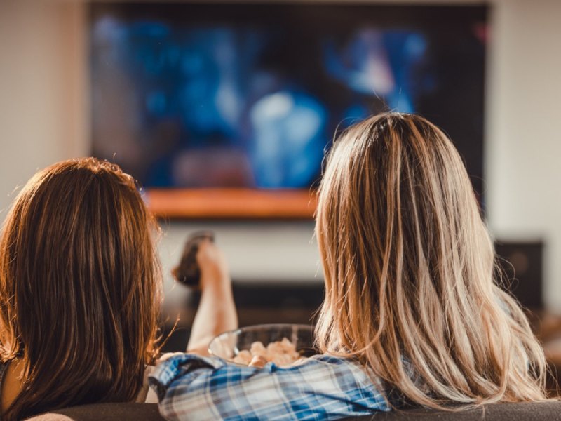 Zwei Personen sitzen auf dem Sofa vor einem Fernseher.