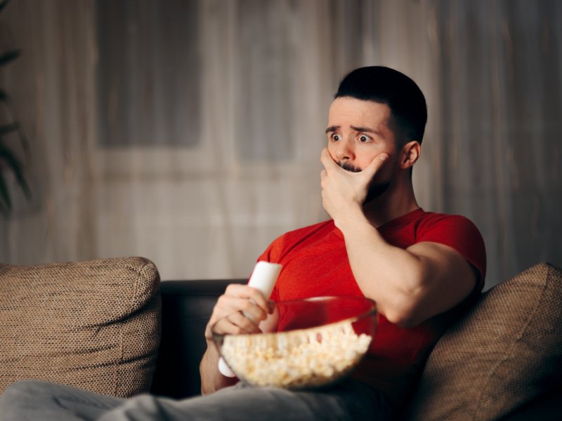 Man schaut einen Film und sitzt dabei auf dem Sofa. Er hat eine Schüssel Popcorn auf dem Schoß und ist erschrocken.