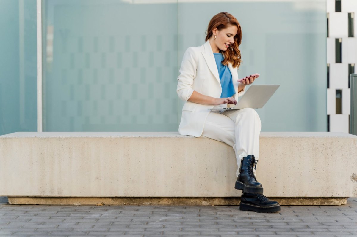 Frau im weißen Anzug sitzt auf einem Stein und nutzt sowohl Laptop als auch Handy.