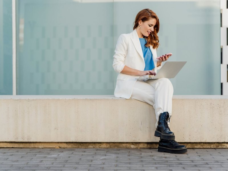 Frau im weißen Anzug sitzt auf einem Stein und nutzt sowohl Laptop als auch Handy.