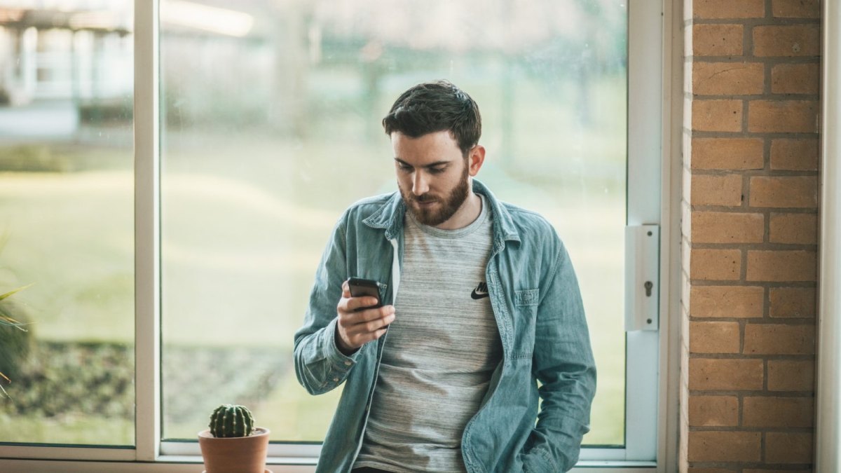 Mann steht vor einem Fenster und schaut auf sein Smartphone.