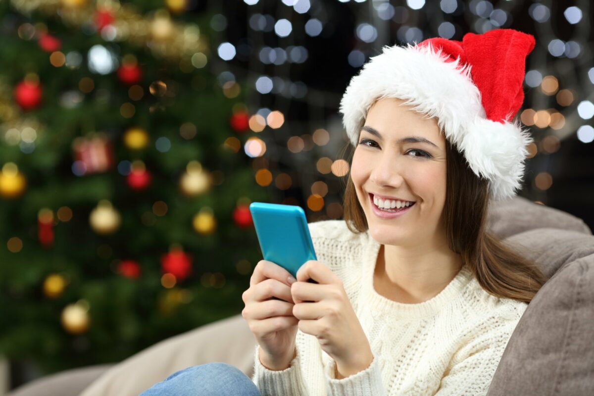 Frau sitzt mit ihrem Handy in einem Weihnachtssetting auf der Couch und guckt in die Kamera.