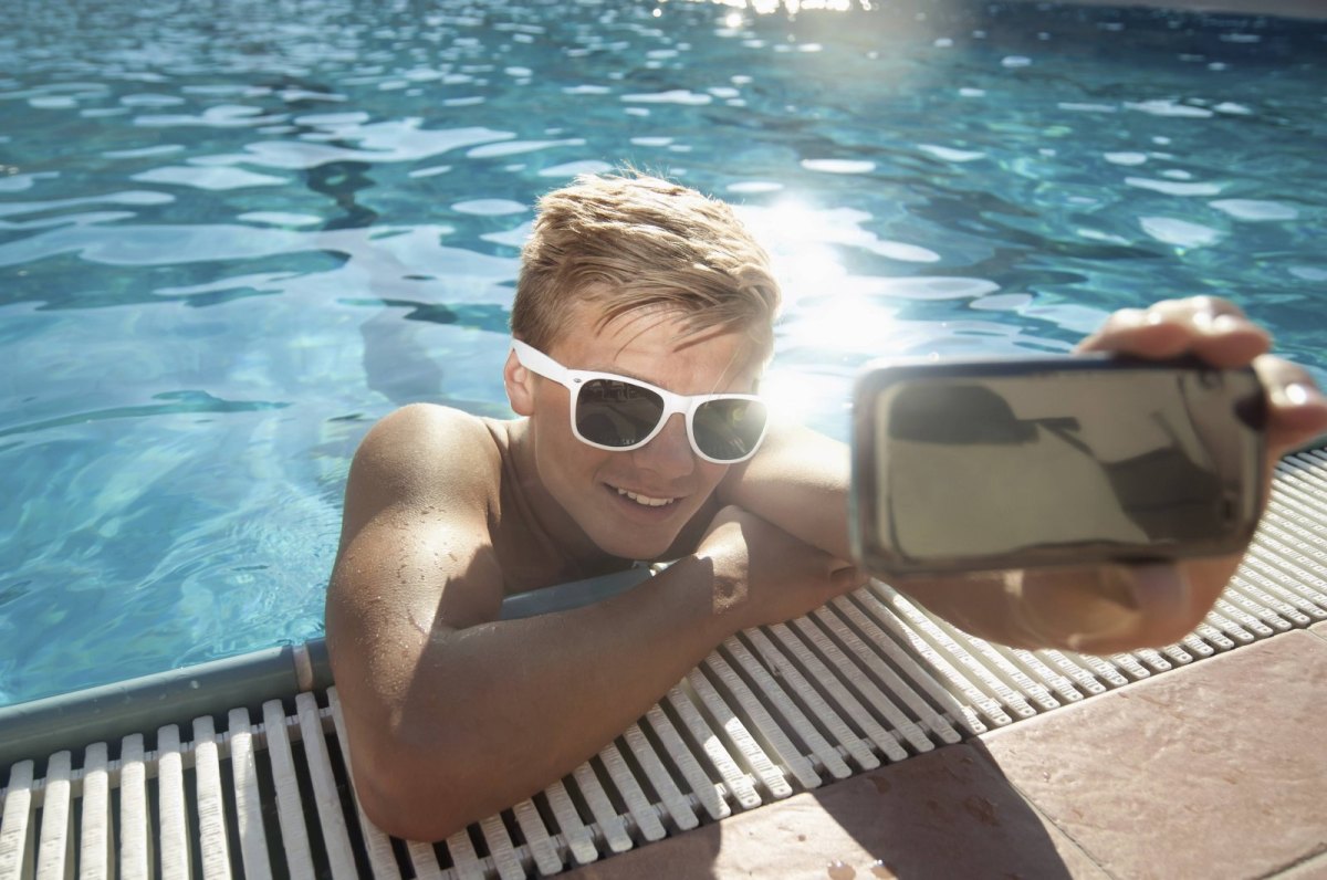 Ein junger Mann mit Sonnenbrille lehnt am Schwimmbeckenrand und macht ein Selfie.