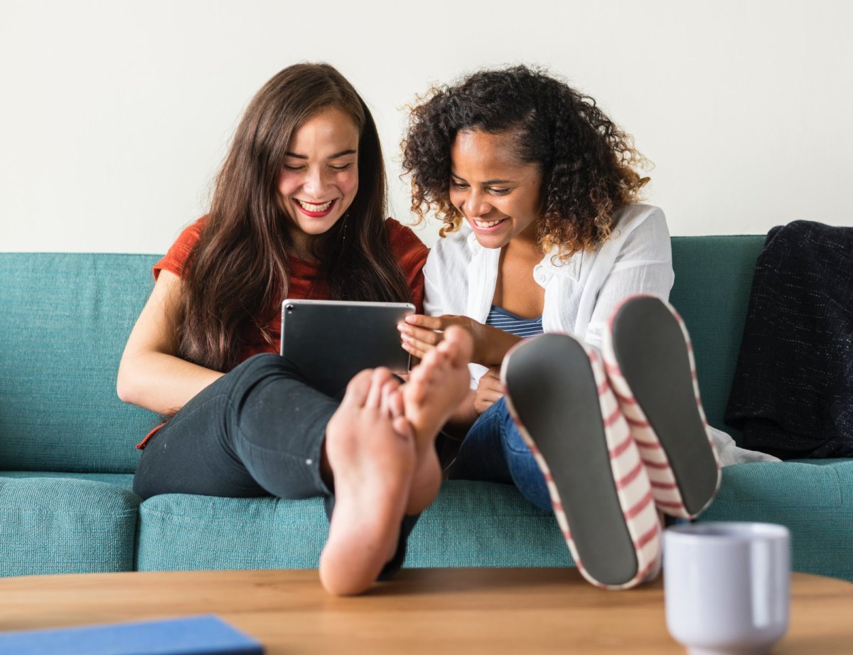 Zwei junge Frauen sitzen auf einer Couch und schauen amüsiert auf ein Tablet.