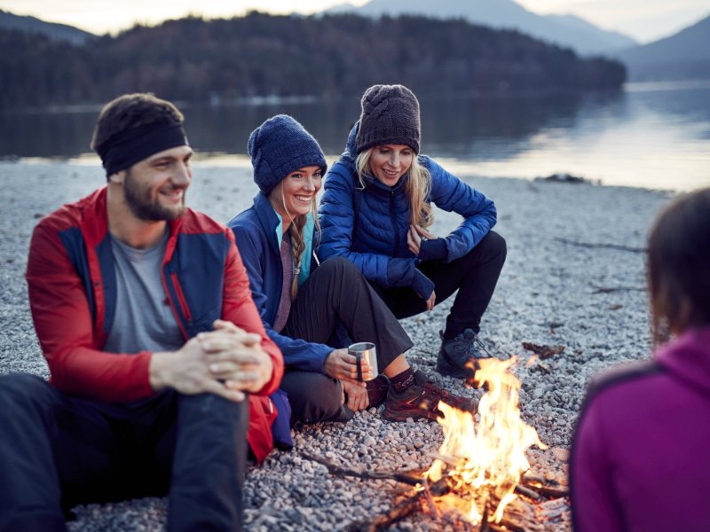 Drei Personen sitzen an einem Lagerfeuer vor einem See