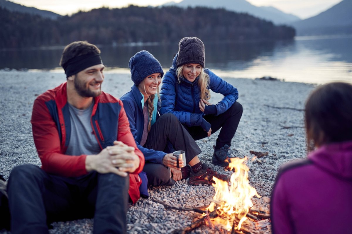 Drei Personen sitzen an einem Lagerfeuer vor einem See