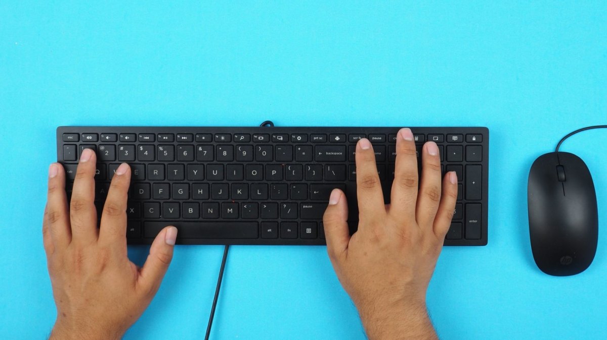 Eine schwarze Tastatur und eine schwarze PC-Maus auf einem hellblauen Untergrund. Zwei Hände tippen auf der PC-Tastatur.