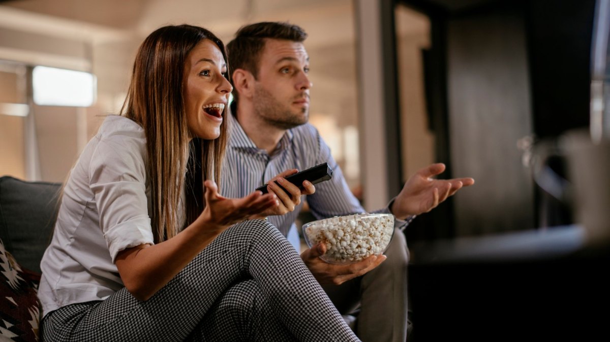 Paar sitzt überrascht und glücklich und mit einer großen Schüssel Popcorn vor dem Fernseher.