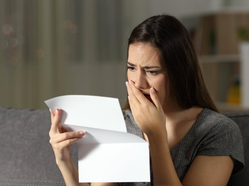 Frau hält ein Blatt Papier in der Hand und schaut geschockt drauf.