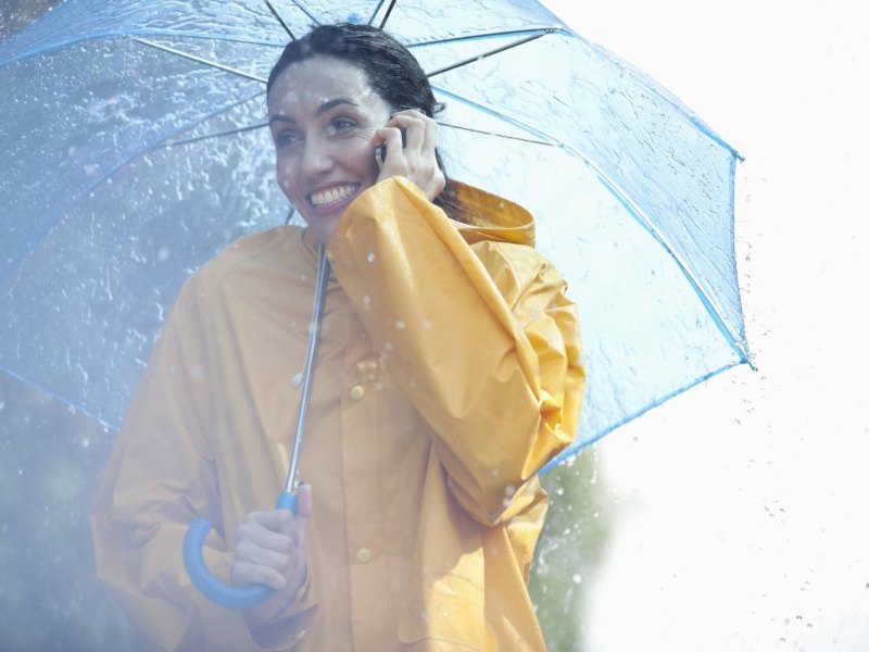 Frau mit Regenschirm und -jacke telefoniert mit ihrem Handy.