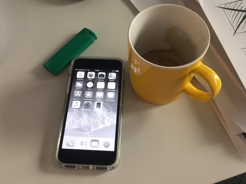 Ein iPhone mit schwarz-weißem Bildschirm liegt neben einer gelben Tasse und einem grünen Feuerzeug auf dem Tisch.