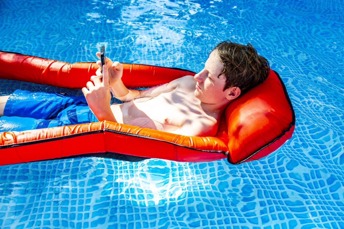Ein Junge liegt auf einer Luftmatratze in einem Pool und spielt am Handy.