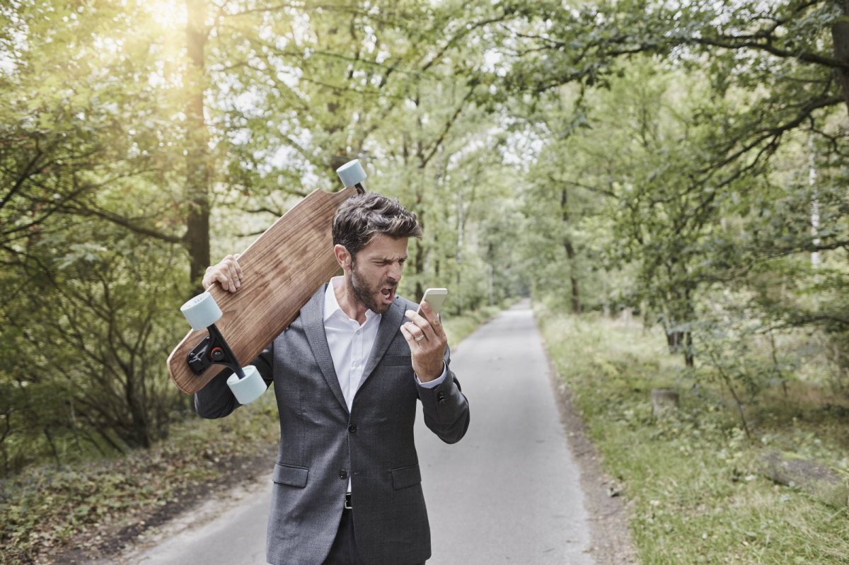 Ein Mann läuft mit einem Longboard durch den Wald und schreit sein Smartphone an.