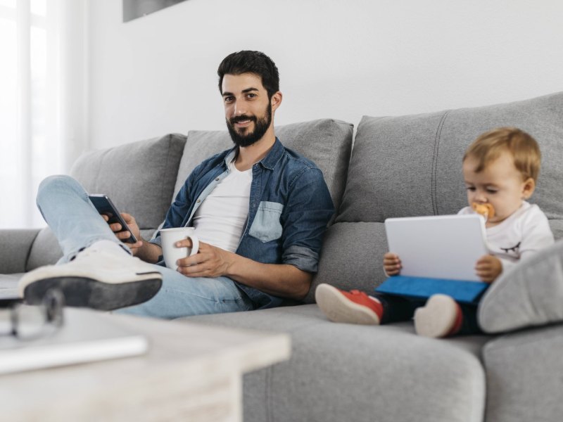 Ein Mann und ein Baby sitzen auf der Couch und schauen auf Tablet/Smartphone.