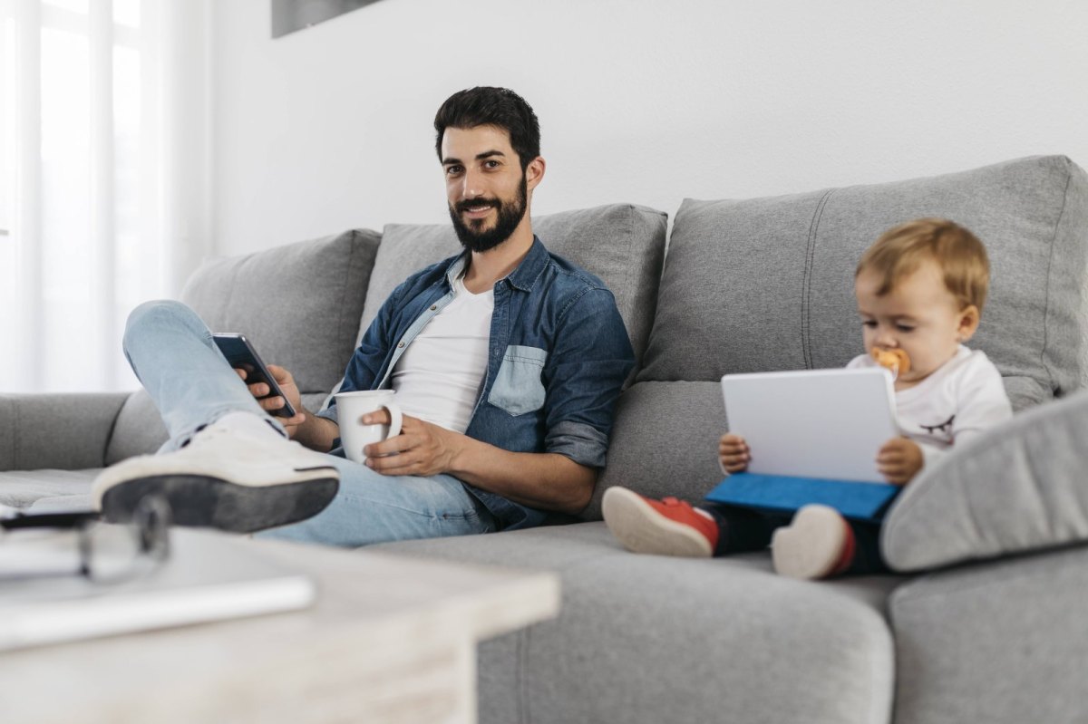 Ein Mann und ein Baby sitzen auf der Couch und schauen auf Tablet/Smartphone.