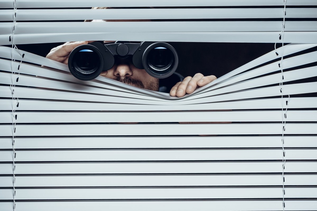 Mann schaut mit einem Fernglas durch eine Jalousie.