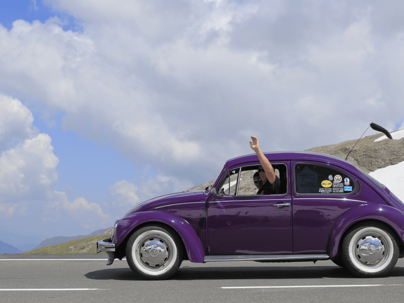 VW Käfer in lila mit winkendem Fahrer