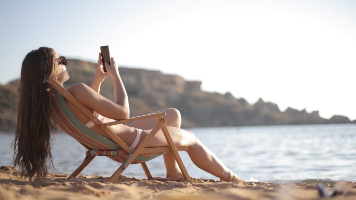 Eine Frau liegt am Strand in einer Liege und schaut auf ihr Smartphone.