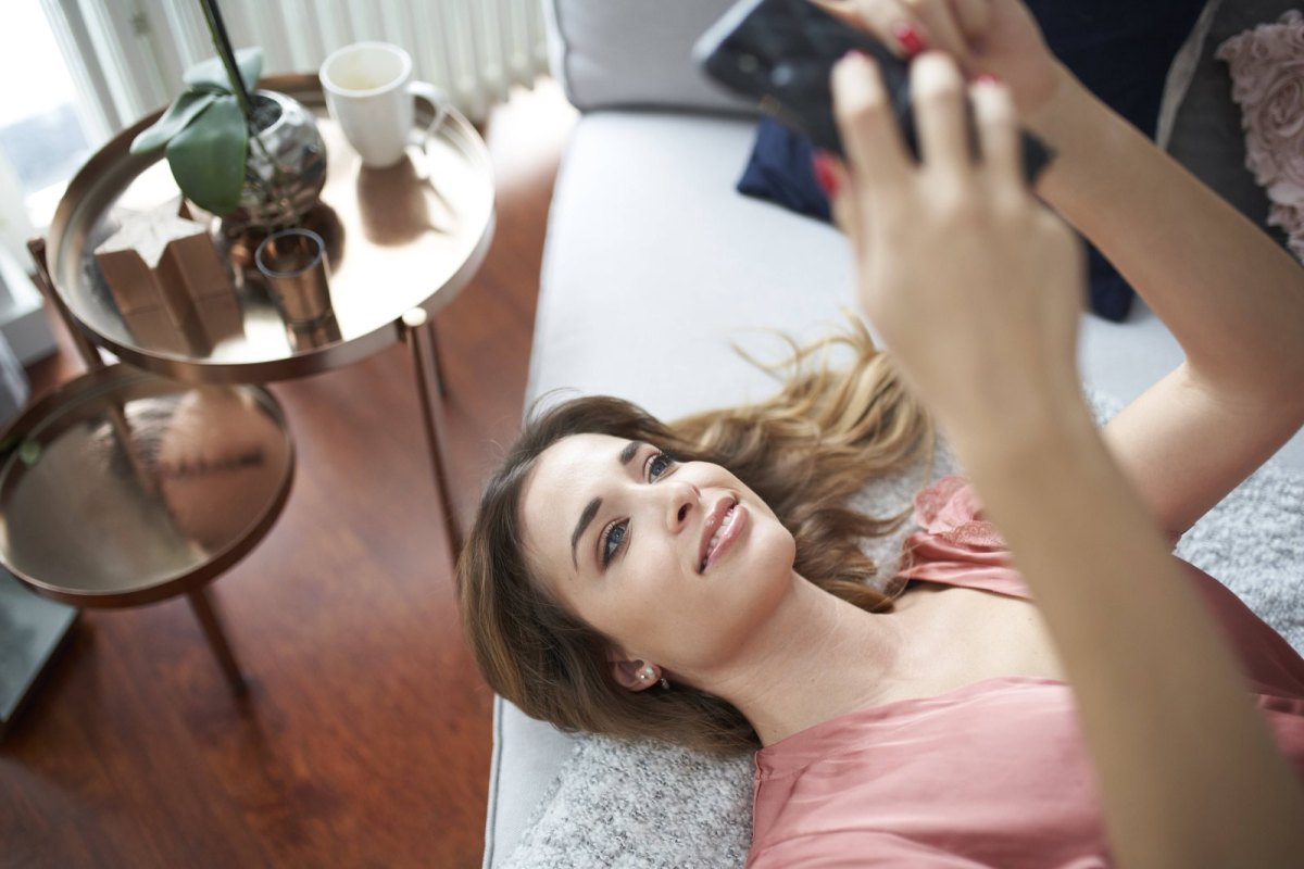 Frau liegt lächelnd mit ihrem Smartphone in der Hand auf einem Bett