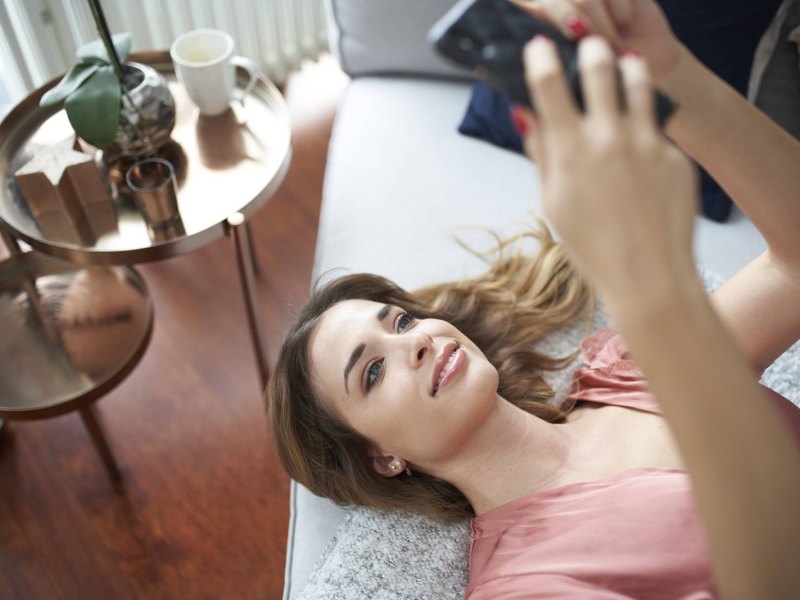 Frau liegt lächelnd mit ihrem Smartphone in der Hand auf einem Bett