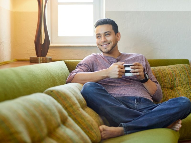 Mann sitzt gemütlich und lächelnd mit einer Tasse in der Hand auf dem Sofa.