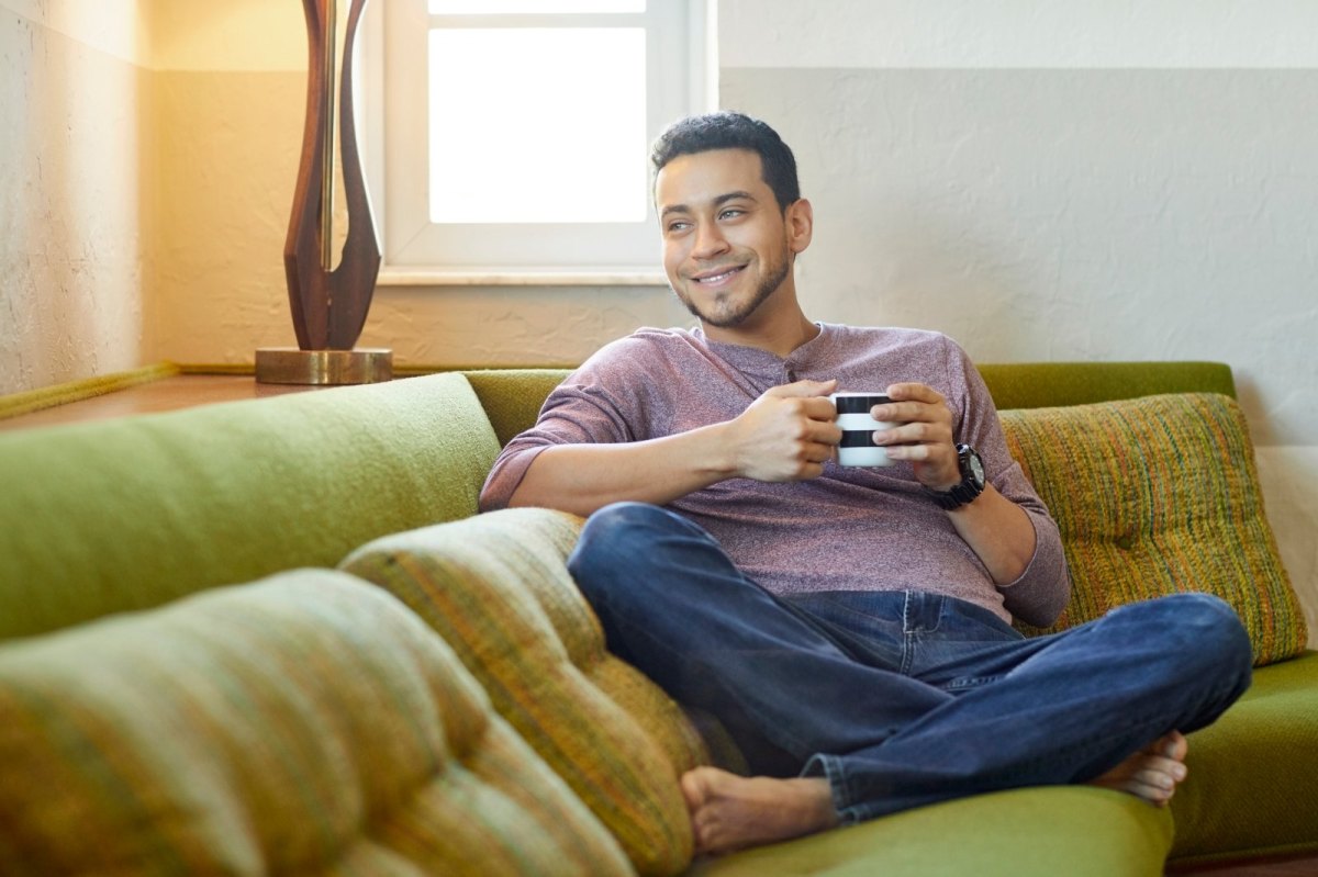 Mann sitzt gemütlich und lächelnd mit einer Tasse in der Hand auf dem Sofa.