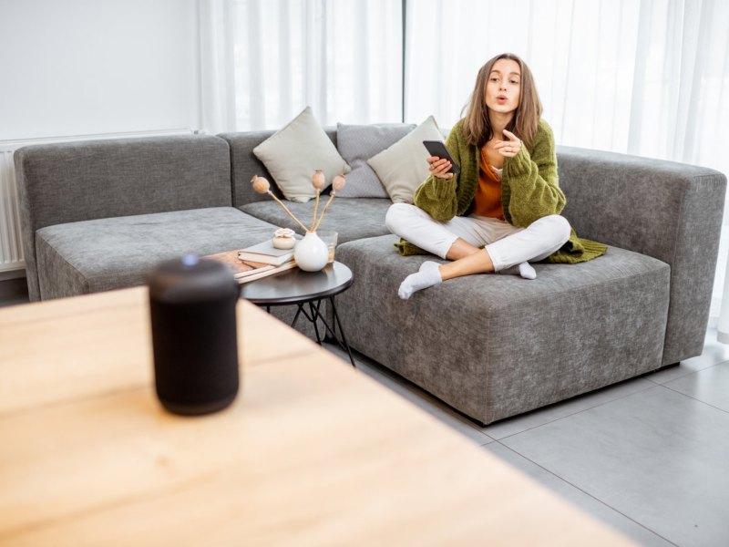 Eine Frau sitzt auf dem Sofa und befiehlt ihrem Heimassistenten etwas.