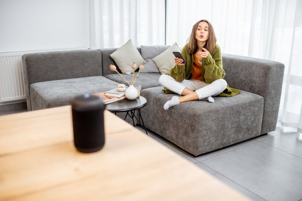 Eine Frau sitzt auf dem Sofa und befiehlt ihrem Heimassistenten etwas.