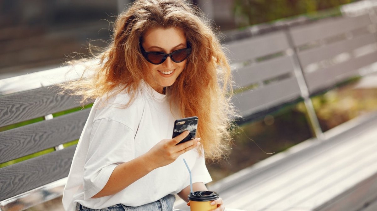 Eine Frau auf einer Bank schaut auf ihr Smartphone und lächelt.