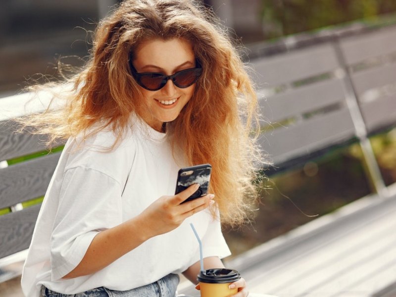 Eine Frau auf einer Bank schaut auf ihr Smartphone und lächelt.