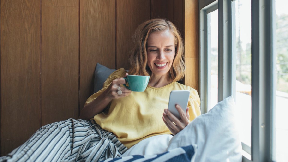 Frau sitzt mit Kaffee und einem Handy in der Hand gemütlich am Fenster.