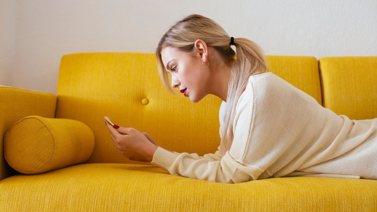 Frau mit Handy auf einem gelben Sofa.