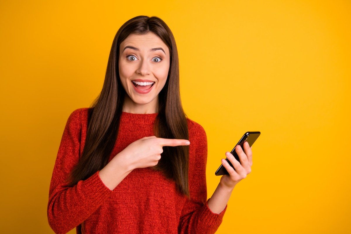 Frau zeigt frühlich mit dem Finger auf ihr Handy.