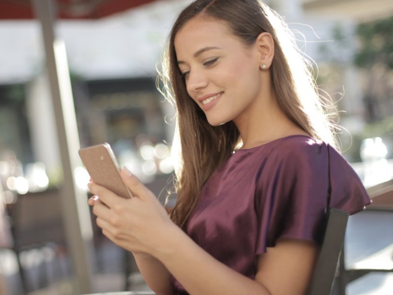 Eine Frau sitzt an einem Tisch und schaut lächelnd auf ihr Smartphone