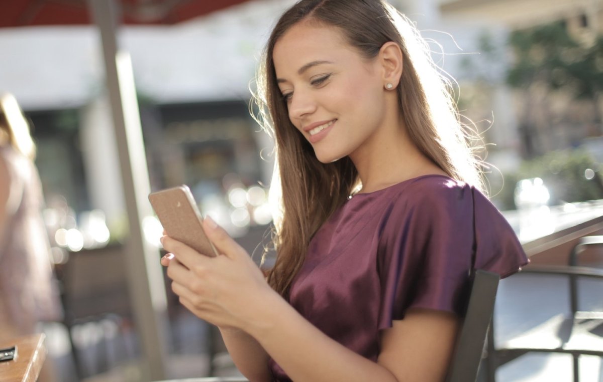 Eine Frau sitzt an einem Tisch und schaut lächelnd auf ihr Smartphone