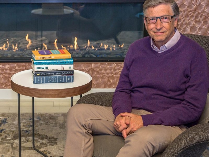 Bill Gates mit Büchern vorm Kamin.