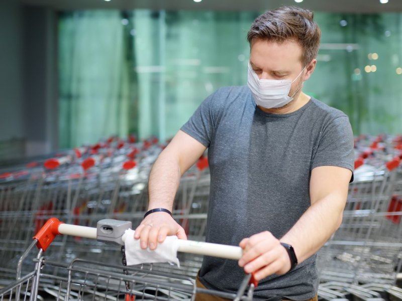 Mann mit Maske desinfiziert einen Einkaufswagen.