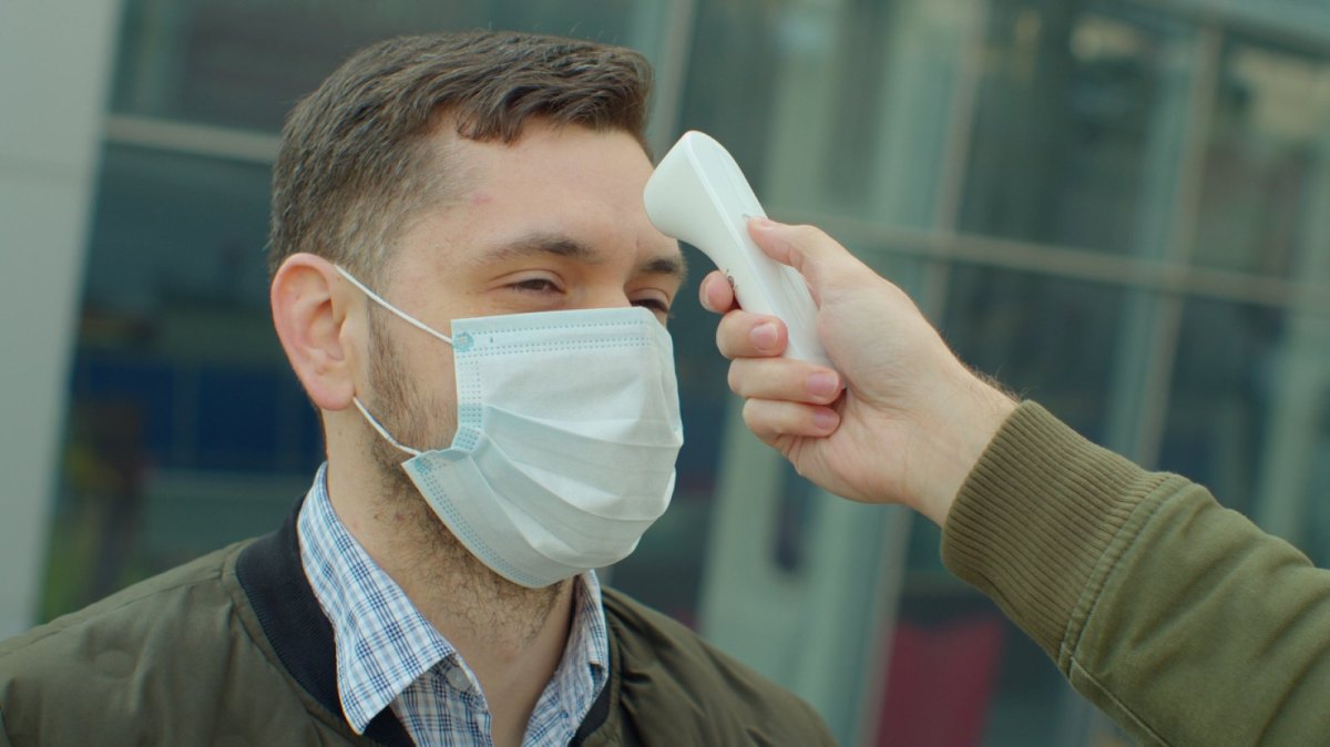 Mann mit Maske bekommt Fieberthermometer an die Stirn gehalten