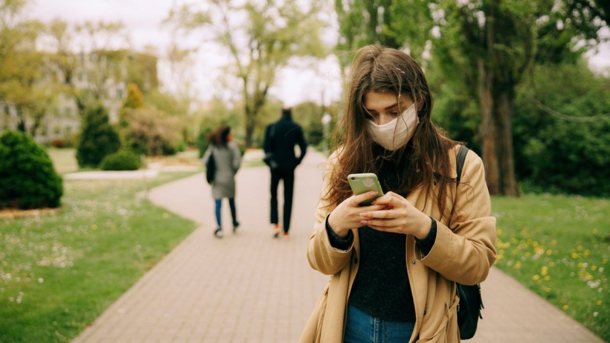 Eine Frau mit Mund-Nasen-Schutz-Maske schaut auf ihr Smartphone.