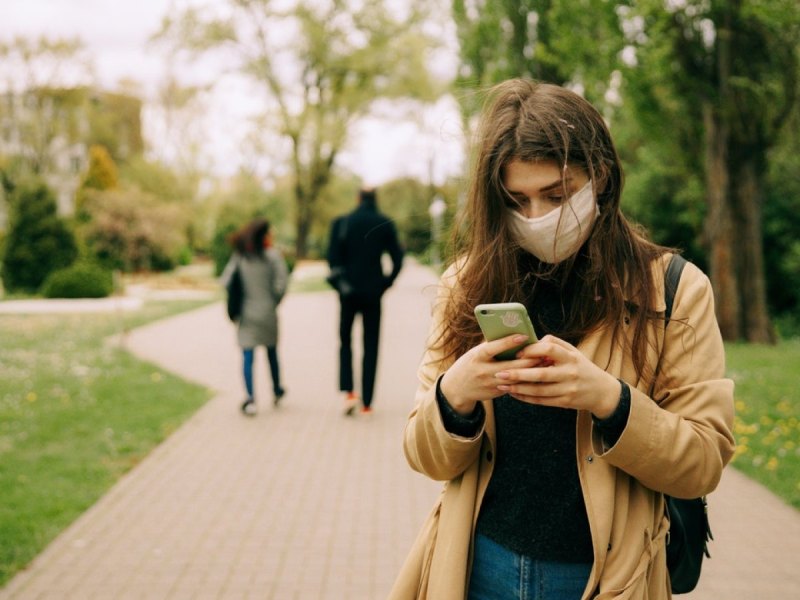 Eine Frau mit Mund-Nasen-Schutz-Maske schaut auf ihr Smartphone.