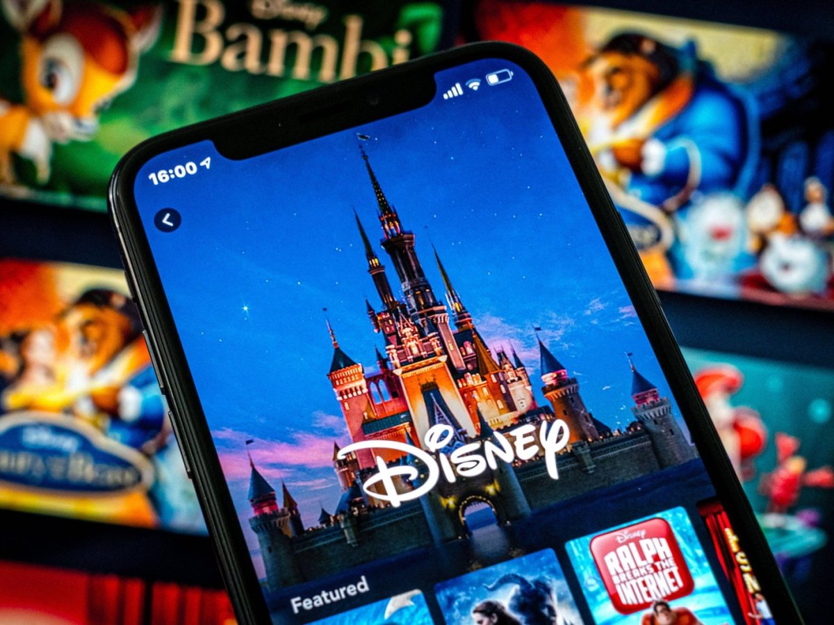Handy mit dem Disney-Logo und dem Disney-Schloss. Im Hintergrund verschwommen: Filmcover von Bambi